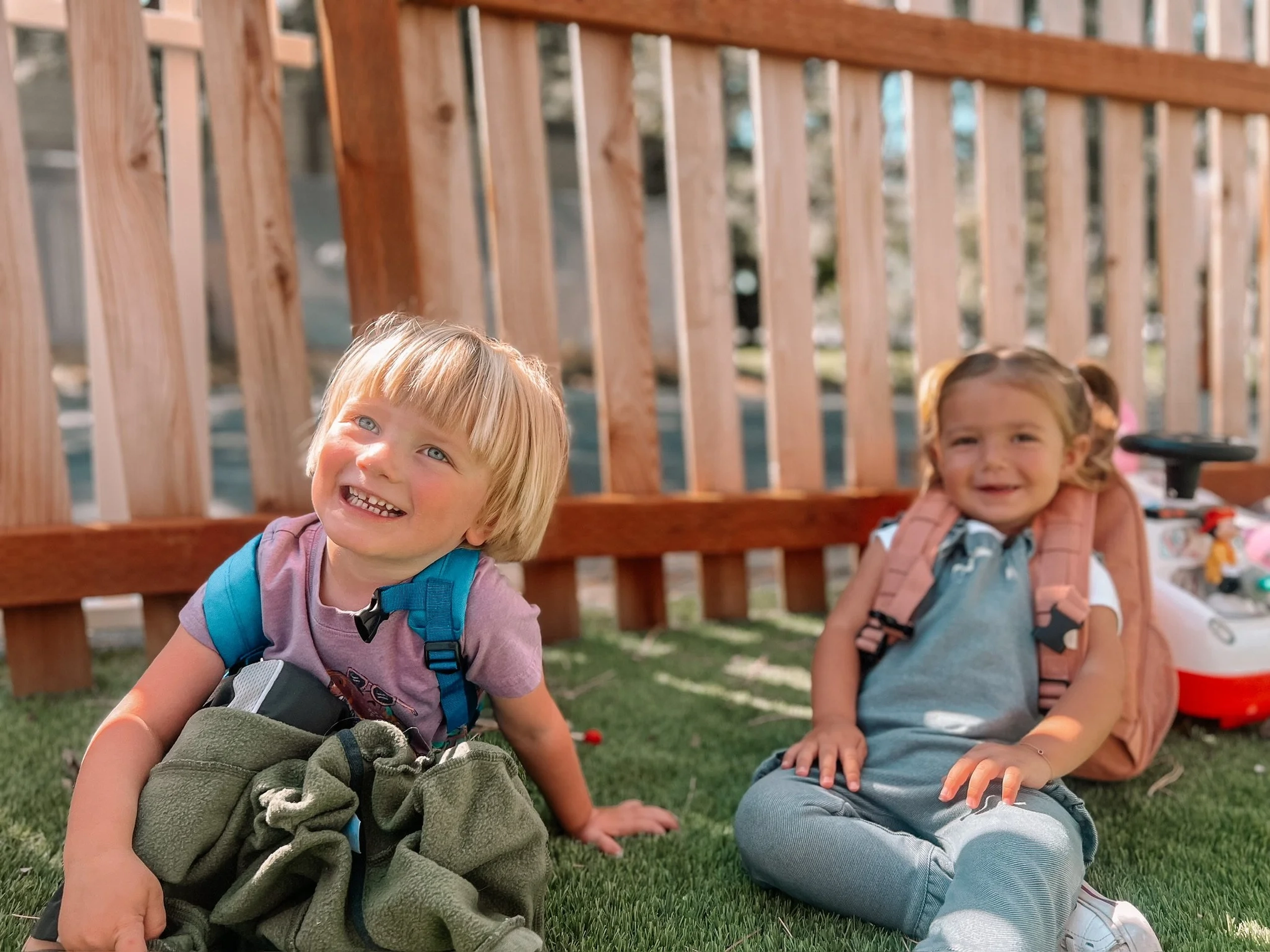 Two children with backpacks sitting on the grass