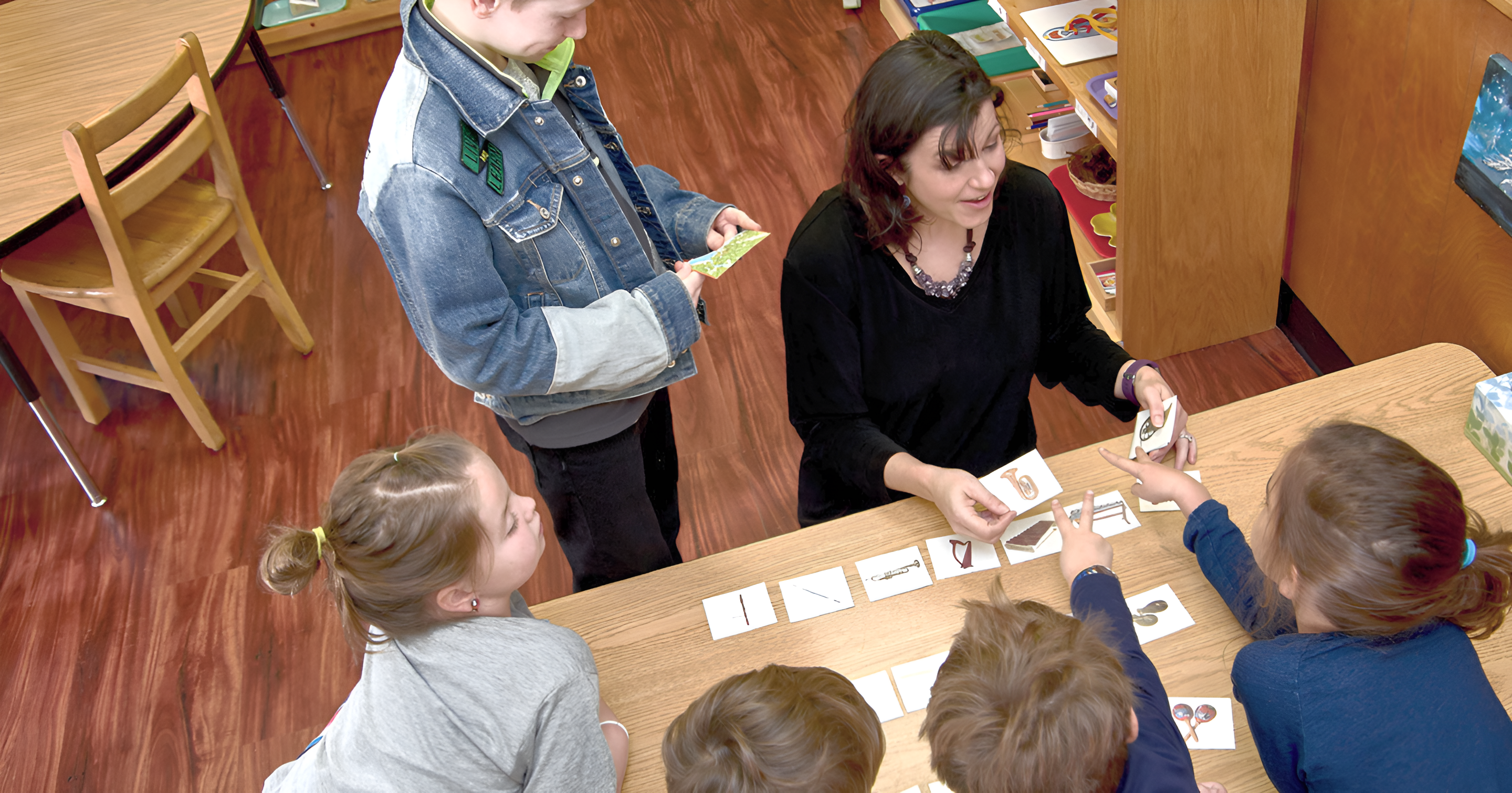 A Montessori guide showing picture cards to five students in a classroom.